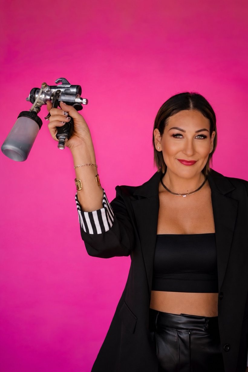 Ashlee Bivins holding an airbrush spray tan gun against a hot pink wall, wearing a black blazer, black top and black leather pants